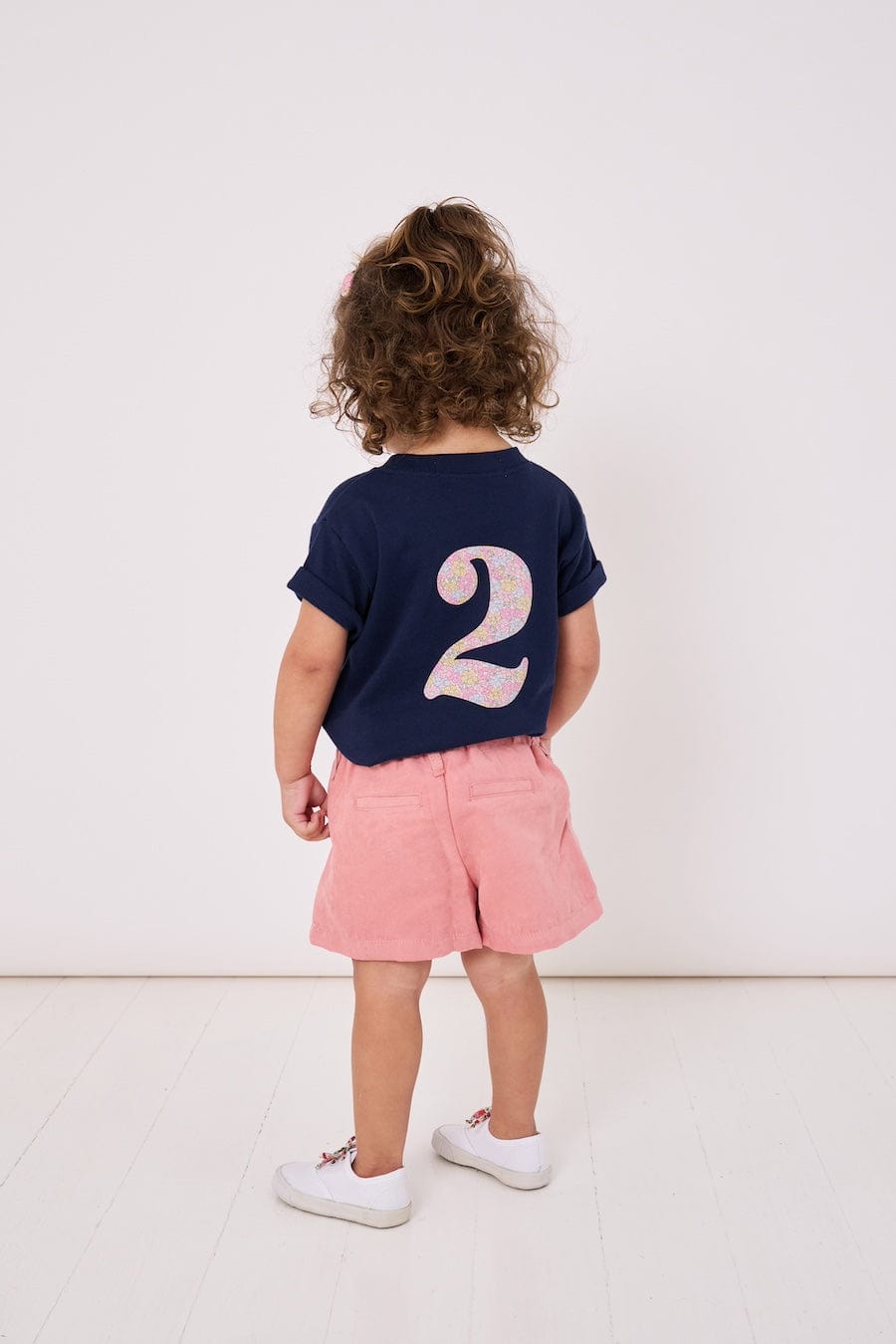 A 2 year old girl wearing a kids cotton t-shirt with a Liberty print number 2 on the back.