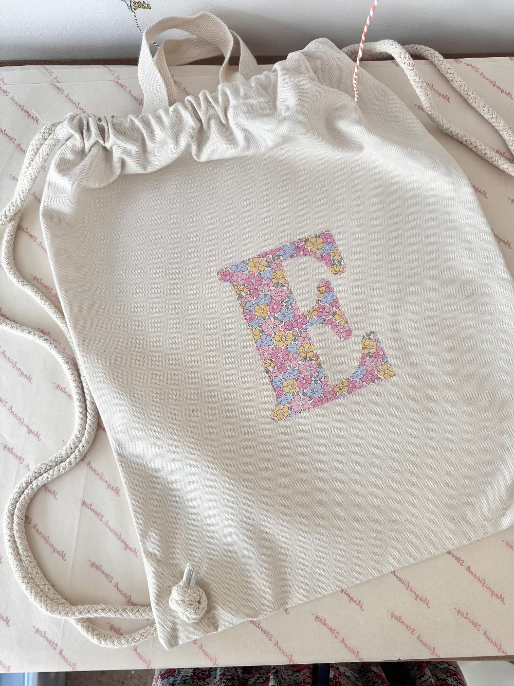 A cream canvas drawstring backpack with a personalised Liberty floral initial sewn on the front