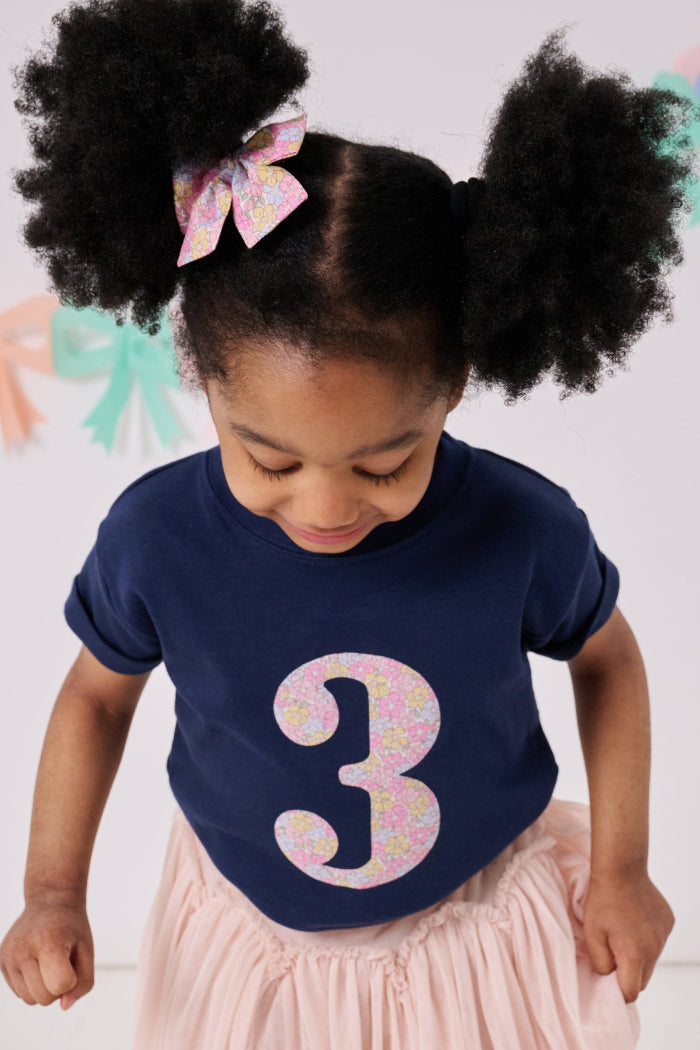 A 3 year old birthday girl wearing a navy cotton kids birthday t-shirt with the  number 3 in Sadie Rose pink floral Liberty print on the front.