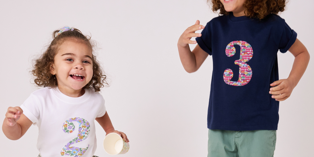 Two young children wearing matching graphic t-shirts with colorful numbers on a light gray studio background. The child on the left wears a white t-shirt with a pink and multicolor number 2, and the child on the right wears a navy blue t-shirt with a pink and multicolor number 3.