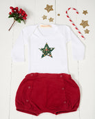 A white long-sleeve baby bodysuit with a green star-shaped patch featuring a festive Liberty Elves print featuring snowdrops, holly, elves and stars, laid flat on a green patterned fabric background. pictured with a candy cane, stars and berries.