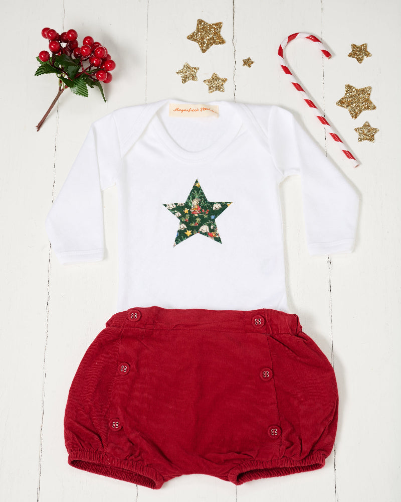 A white long-sleeve baby bodysuit with a green star-shaped patch featuring a festive Liberty Elves print featuring snowdrops, holly, elves and stars, laid flat on a green patterned fabric background. pictured with a candy cane, stars and berries.