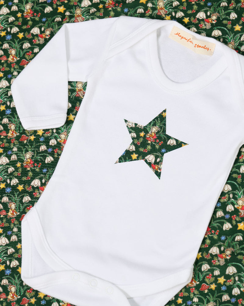 A white long-sleeve baby bodysuit with a green star-shaped patch featuring a festive Liberty Elves print featuring snowdrops, holly, elves and stars, laid flat on a green patterned fabric background. 