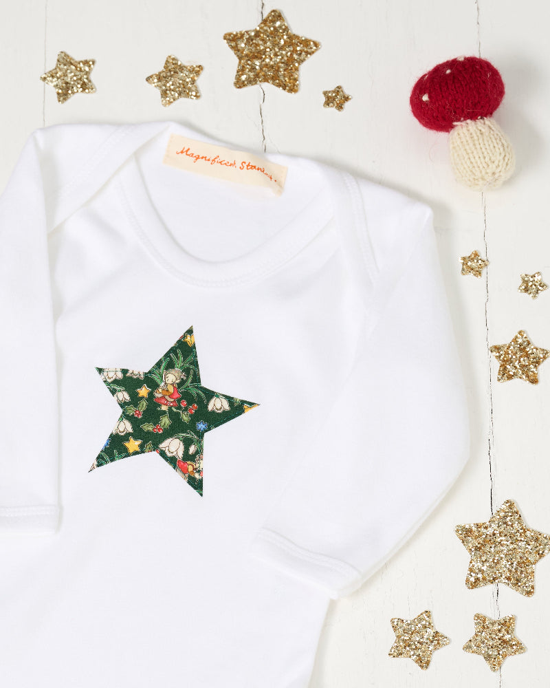 A white long-sleeve baby baby grow  with a green star-shaped patch featuring a festive Liberty Elves print featuring snowdrops, holly, elves and stars, laid flat on a green patterned fabric background. 