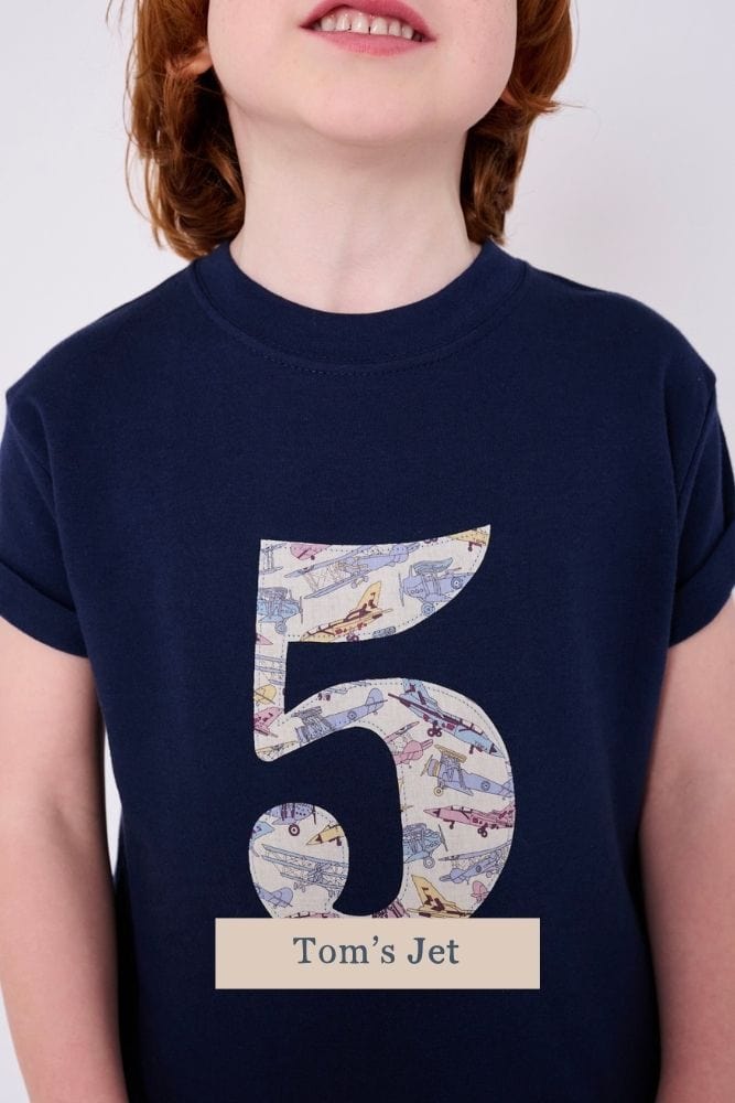 a 5 year old boy wearing a navy birthday t-shirt with a number 5 cut from Tom's Jet plane liberty print on the front