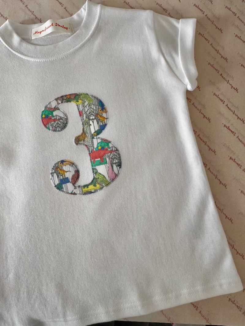 A white short-sleeve baby tee with a colorful patchwork-style number 3 printed on the chest, laid flat on patterned paper. The brand label inside the collar reads 'Magical Dresses'.