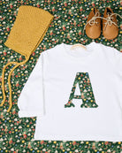 A styled flat-lay of a white long-sleeve baby t-shirt featuring a large letter 'A' sewn on the chest in a green floral Liberty elves pattern.
