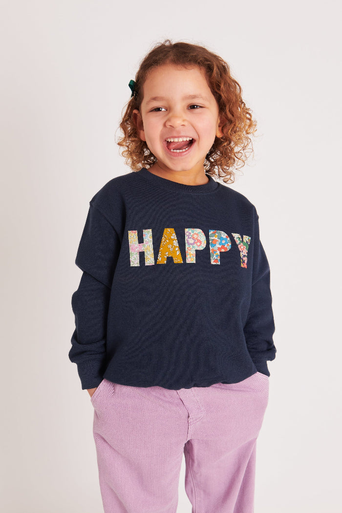 A 5 year old girl wearing a Magnificent Stanley navy sweatshirt with the word HAPPY Mixed Liberty Print sewn onto the front
