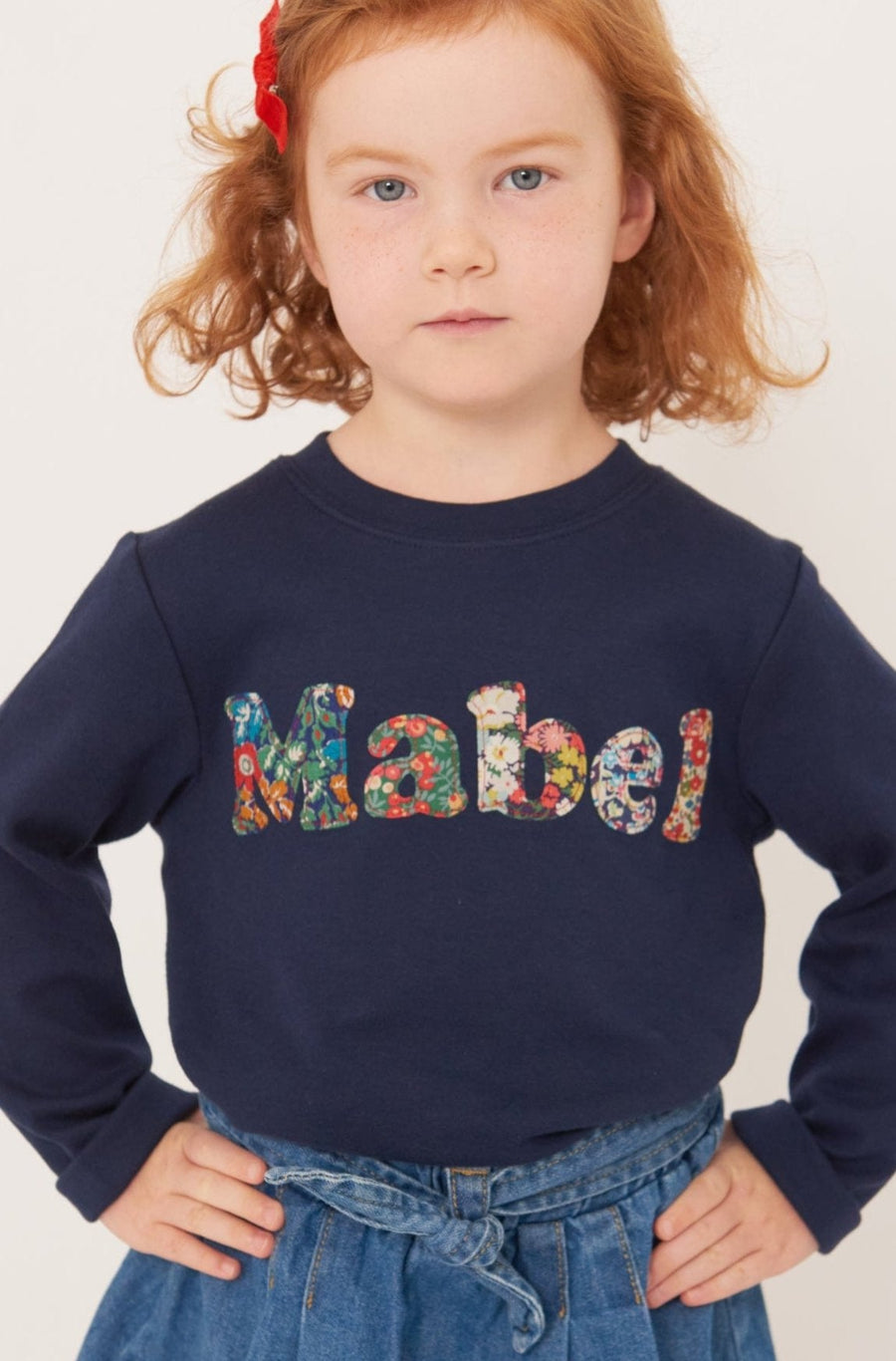A 4 year old girl wearing a navy t-shirt with her name sewn on in mixed Liberty print lowercase letters.