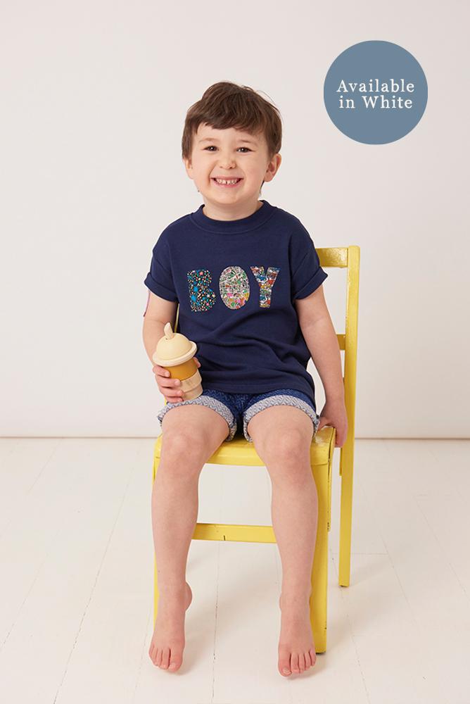 A little boy wearing a navy t-shirt with a name on in mixed Liberty fabrics.