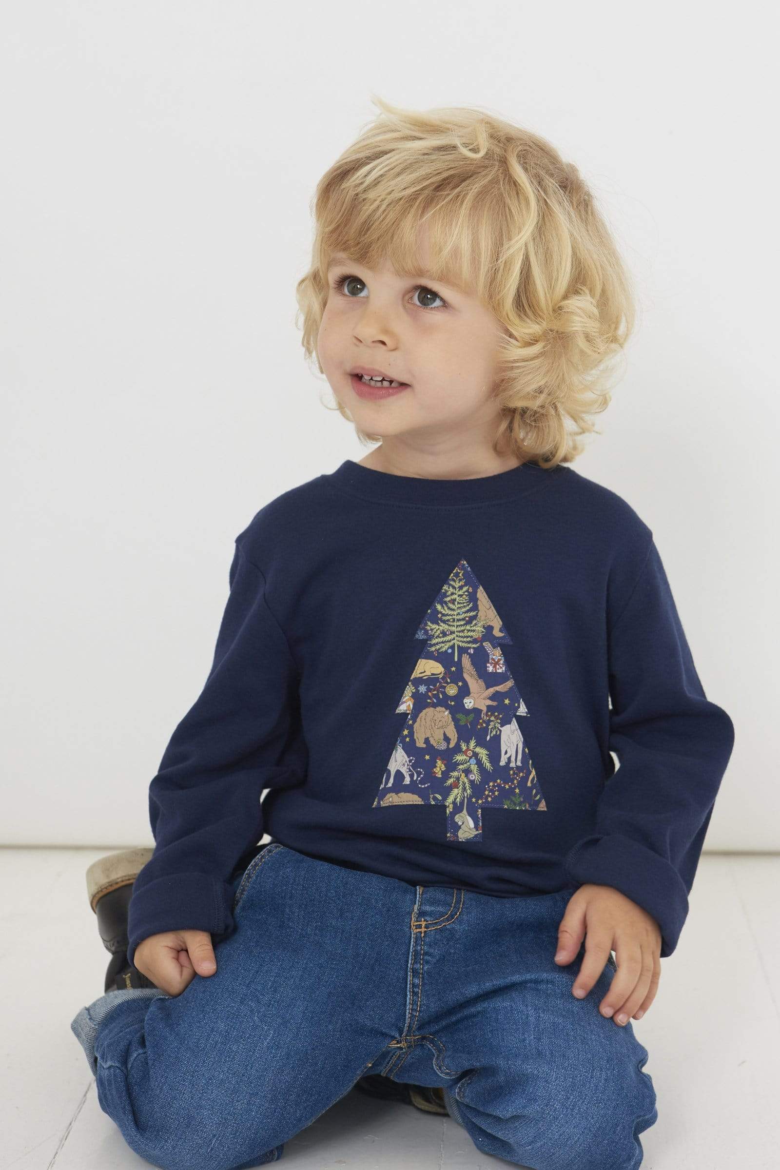 A 3 year old boy wearing a navy long sleeve Christmas t-shirt with a Christmas Tree on the front made from Liberty Christmas print .