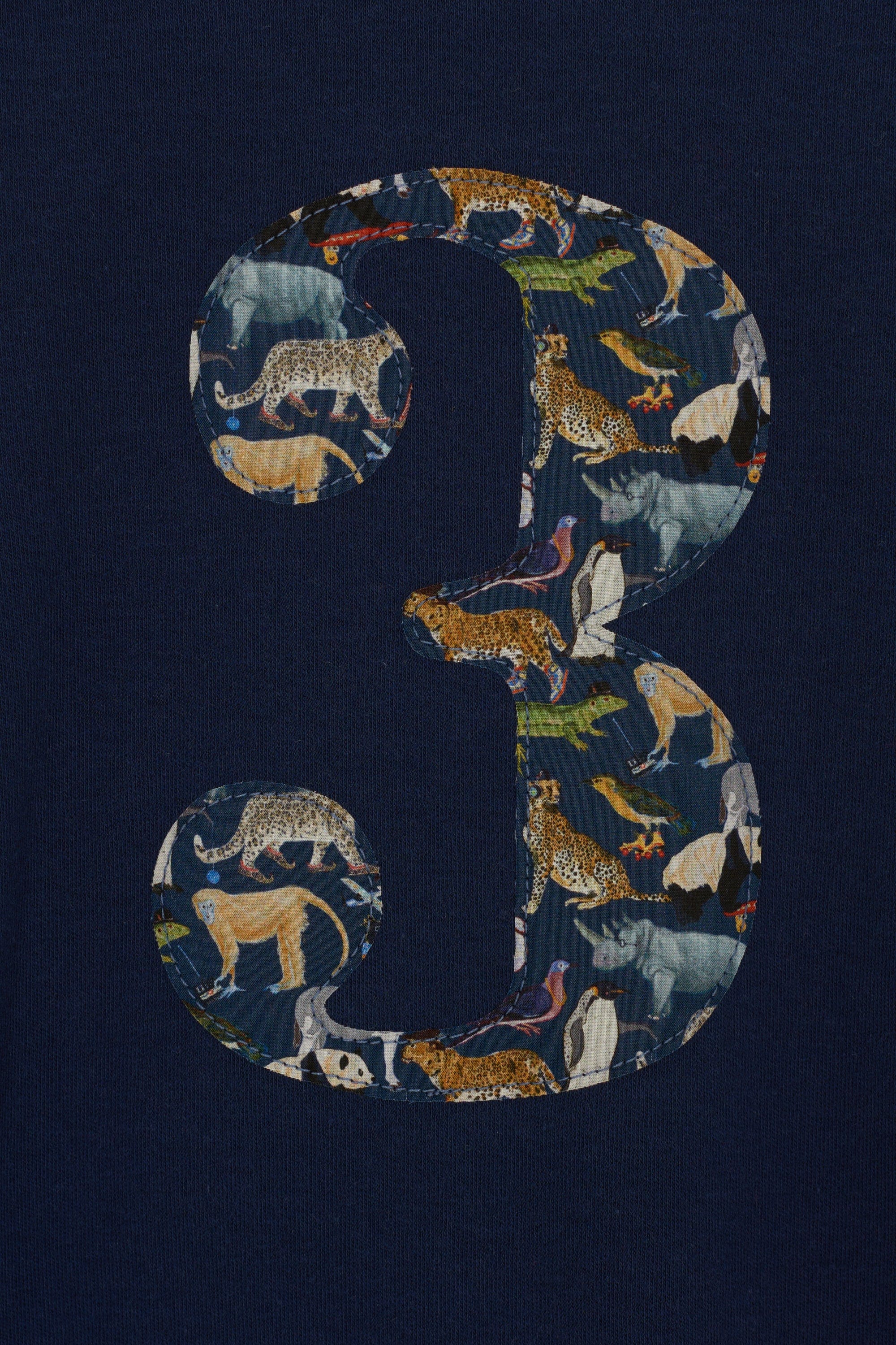 A 3 year old boy wearing a navy long sleeve t-shirt with a  number 3 on the front on in Liberty Quey Zoo animal print fabric. A close up of the appliqué stitching.