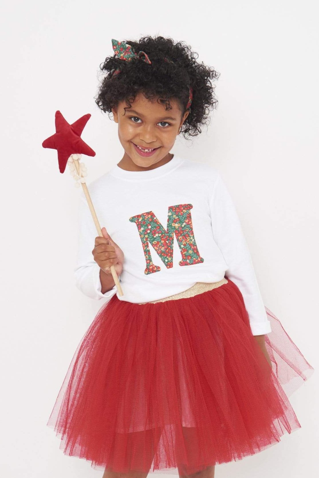 A 5 year old girl wearing a personalised white t-shirt with a her initial on the front made from Liberty Glitter Wiltshire berry fabric.
