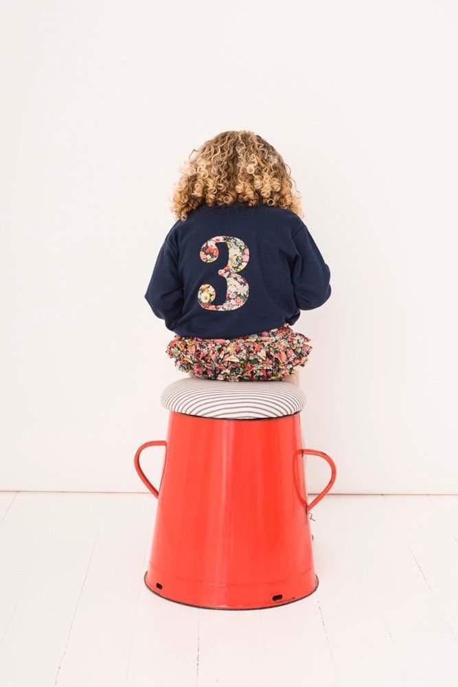 A 3 year old girl wearing a navy cotton tee with a large number 3 on the back in floral Liberty fabric.